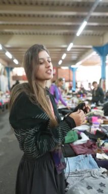 Genç bir kadın renkli giysileri zevkle keşfediyor. Canlı bir pazar yerinde, çeşitli ürünlerle dolu.