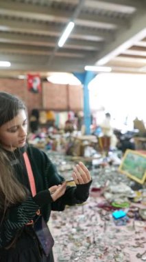 Meraklı genç bir kız, hareketli bit pazarındaki canlı ve eklektik eşyalar arasında bir barış sembolünü dikkatle inceliyor.