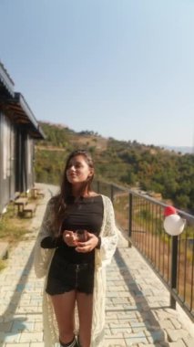 Bir kadın açık havada güneşli bir günün tadını çıkarırken güzel, manzaralı bir arka plana karşı ferahlatıcı bir içecek tutar.