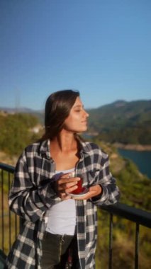 Genç bir kadın balkonda dinlenmek için zaman ayırıyor. Etrafı göz kamaştırıcı doğal manzarayla çevriliyken içkisini yudumluyor.