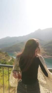 Genç bir kadın sakin bir manzaranın yanında zarif bir şekilde duruyor, güzel bir ortamda ferahlatıcı bir içecek tutuyor.