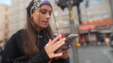 Canlı bir şehir ortamında, genç bir kadın canlı şehir hayatı ile çevriliyken akıllı telefonuyla meşgul olur.