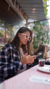 Moda bir kadın açık hava kahvesinde akıllı telefonuna odaklanırken kahvesini içiyor.