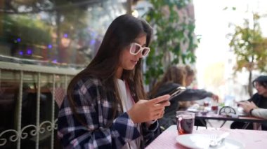 Açık bir kafede akıllı telefonuna dalmış şık bir genç kadın kahvesinin tadını çıkarıyor.