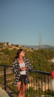 Şık bir kadın balkonda duruyor, güzel bir günbatımı manzarasının keyfini çıkarırken doğa güzelliği ile çevrili.