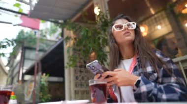 Tarz sahibi bir kadın kafede oturur, telefonunu kontrol eder ve açık havada çayını yudumlar.