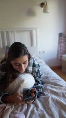 Genç bir kadın sıcak ışıklarla dolu sıcak bir odada, akıllı telefonuna derinden odaklanmış bir şekilde yatağında oturuyor.