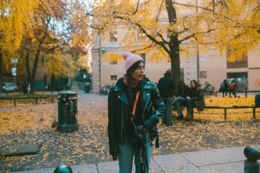 Şık deri ceketli ve şık bereli genç bir adam renkli sonbahar yapraklarıyla güzel bir parkta duruyor.