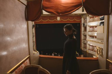 Siyah elbiseli zarif giyinmiş bir kadın, düşünceli bir şekilde tiyatro kutusundan bakıyor, zamansız sanatın vücut bulmuş hali.