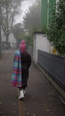 Issız bir figür, canlı ve renkli bir battaniyeye sarılmış, sisli bir sokakta sakin bir şekilde geziniyor.