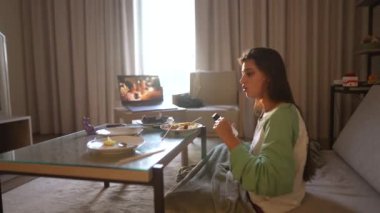 Bir kadın rahat odasında rahatlar, TV izlerken atıştırmalıkların tadını çıkarır, evde huzurlu bir yaşam tarzı yakalar.