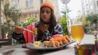 Çevresi yeşillik ve şehir atmosferiyle çevrili açık bir kafede lezzetli suşinin tadını çıkaran bir insan.