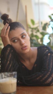 Yeşillik ve içecekle çevrili bir kafede düşüncelere dalmış genç bir kadın.