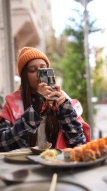 Genç bir kadın, açık hava ortamında telefonuyla ustaca fotoğraf çekerken suşi yemekten zevk alır.