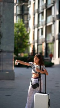 Şık bir genç kadın şık bavuluyla neşeli bir selfie çekiyor. Canlı bir şehir ortamında.