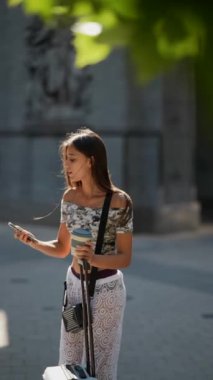 Genç bir kadın şehir ortamında dururken telefonunu kontrol ediyor, bavuluyla biraz zaman geçiriyor.
