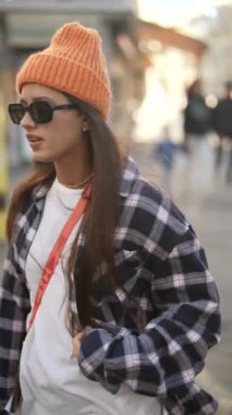 Modayı takip eden genç bir kadın canlı bir caddede ekose gömlek ve turuncu bir bere giyerek yürüyor.