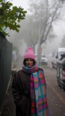 Bir kadın sisli bir sokakta duruyor, renkli bir eşarp ve şapkaya sarılmış, huzurlu bir atmosfer yaratıyor..