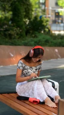 Genç bir kadın, yaratıcılığına tamamen dalmış, kulaklık takmış, açık havada huzur içinde resim çiziyor.