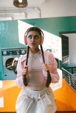 Şık bir çamaşırhanede, modayı takip eden genç bir kadın kulaklık takıyor ve etrafındaki şeylere dalmış bir şekilde rahatça poz veriyor.