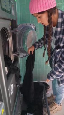 Genç bir kadın, çamaşırhanede bulunan çamaşır makinesine özenle bir demet siyah kıyafet yüklüyor.