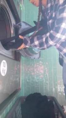 Pembe bereli genç bir kadın çamaşırhanede çamaşır makinesiyle etkileşim kurmakla meşgul.