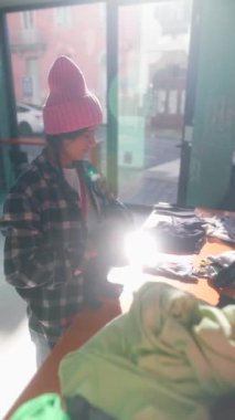 Pembe bereli ve flanel gömlekli biri müşteriler için parlak bir butikte renkli kıyafetler düzenliyor.
