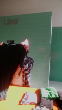 Şık pembe şapkalı bir kişi, parlak, renkli bir çamaşırhane içinde özenle siyah giysileri katlıyor.
