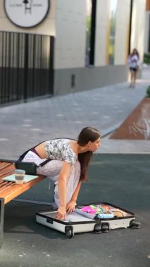 Genç bir kadın bankta oturuyor, renkli kıyafetlerini şık bir kentsel alanda bir bavulda düzenliyor..
