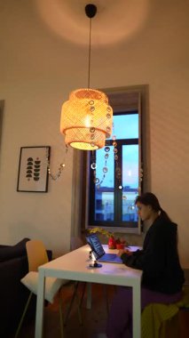 Modern bir stil lambası, odaklanmış bir insan ve parlak doğal ışık içeren davetkar ve sıcak bir çalışma alanı.