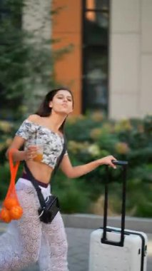 Şık, genç bir kadın, bavulunu arkasından çekerken canlı ve hayat dolu bir şehir manzarasında rahatça yürüyor.