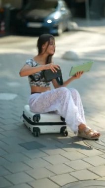 Genç bir kadın kaldırıma çömelmiş, bavulunu hareketli bir şehir ortamında dikkatlice düzeltiyor.