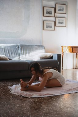 Genç bir kadın yoga rutinini uyguluyor. Parlak, huzurlu bir yaşam alanında bir paspasın üzerinde geriniyor.