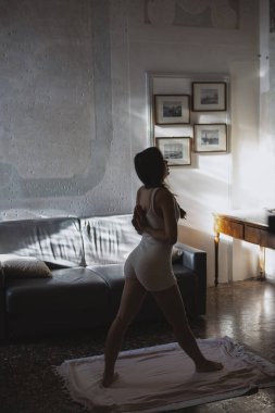 Nazik güneş ışığıyla hafifçe aydınlatılan sakin bir ev ortamında özenle yoga yapan zarif bir figür.