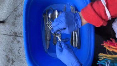 Bir kişi, eldiven giyerken ve çeşitli çatal bıçaklarla dolu mavi bir leğen kullanırken özenle gümüş takımları temizliyor.