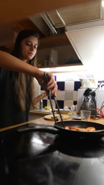 Genç bir kadın yemek pişirme yeteneklerini sergiliyor, malzemelerini maşayla tavada ustalıkla çeviriyor.