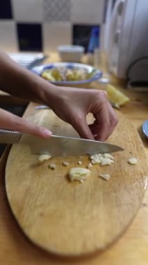 Yakından bakıldığında, mutfakta ahşap bir kesme tahtasıyla taze sarımsağı ustalıkla kesen eller görülüyor.