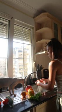 Parlak ve neşeli bir mutfakta, bir kadın taze sebze ve renkli meyveleri yanında bir kediyle yıkıyor.