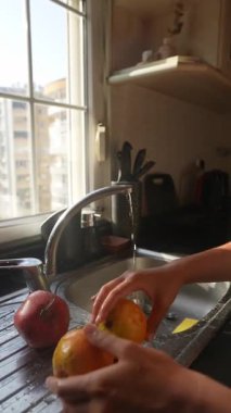 Neşeli bir insan mutfak lavabosunda portakal ve elmaları dikkatlice yıkıyor, sıcak güneş ışığının tadını çıkarıyor.