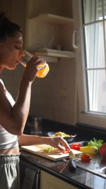 Bir kadın, güneşli ve sıcak mutfağında içkisini yudumlarken bir dizi taze sebzeyi ustalıkla hazırlar.