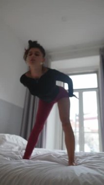 Bir dansçı yatakta akıcı bir poz sergiler, yumuşak doğal ışıkla aydınlatılır, zarafet ve hareket ifade eder.