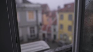 Yağmurlu bir şehir manzarasının biraz bulanık bir görüntüsü bir pencereden görünüyor, güzel renkli binalar sergileniyor.