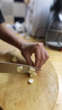 Sıcak bir mutfak ortamında, sağlam bir ahşap kesme tahtasında sarımsak karanfillerini ustalıkla doğrayan bir insan.