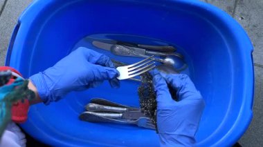 Bir insan, maksimum hijyen için mavi bir leğende gümüş takımları temizlerken etkili bir şekilde eldiven giyer.