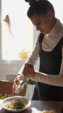 Güneşli bir mutfakta taze bir yemeğin üzerinde peynir rendeleyen bir kadın aşçılık becerilerini ve tutkularını sergiliyor..