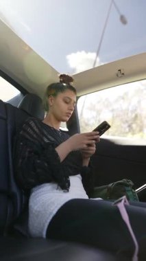 Gündelik kıyafet giymiş genç bir kadın, rahat bir şekilde arabada otururken, dikkatle akıllı telefonuna odaklanır.
