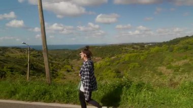 Genç bir kadın, bereketli tepeler ve okyanus manzaralarıyla çevrili, doğa güzelliğini kucaklayan manzaralı bir yolda yürüyor.