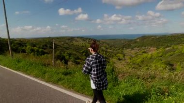 Genç bir kadın, yemyeşil yemyeşil ve çarpıcı mavi bir okyanus zeminiyle süslenmiş güzel bir sahil yolunda yürüyor.
