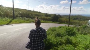 Zarif kareli gömlekli genç bir kadın verimli yeşilliklerle çevrili manzaralı bir yolda rahatça yürüyor.