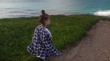 Genç bir kadın yemyeşil ve çarpıcı okyanus manzaralarıyla süslenmiş güzel bir sahil yolunda rahatça yürüyor.